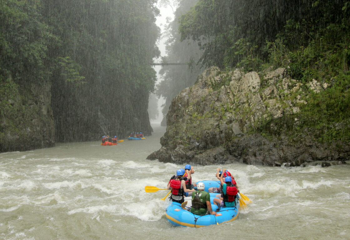 A group of people whitewater rafting in a blue inflatable raft through a narrow river gorge during heavy rain, surrounded by steep, lush green cliffs. Other rafts are visible in the background.