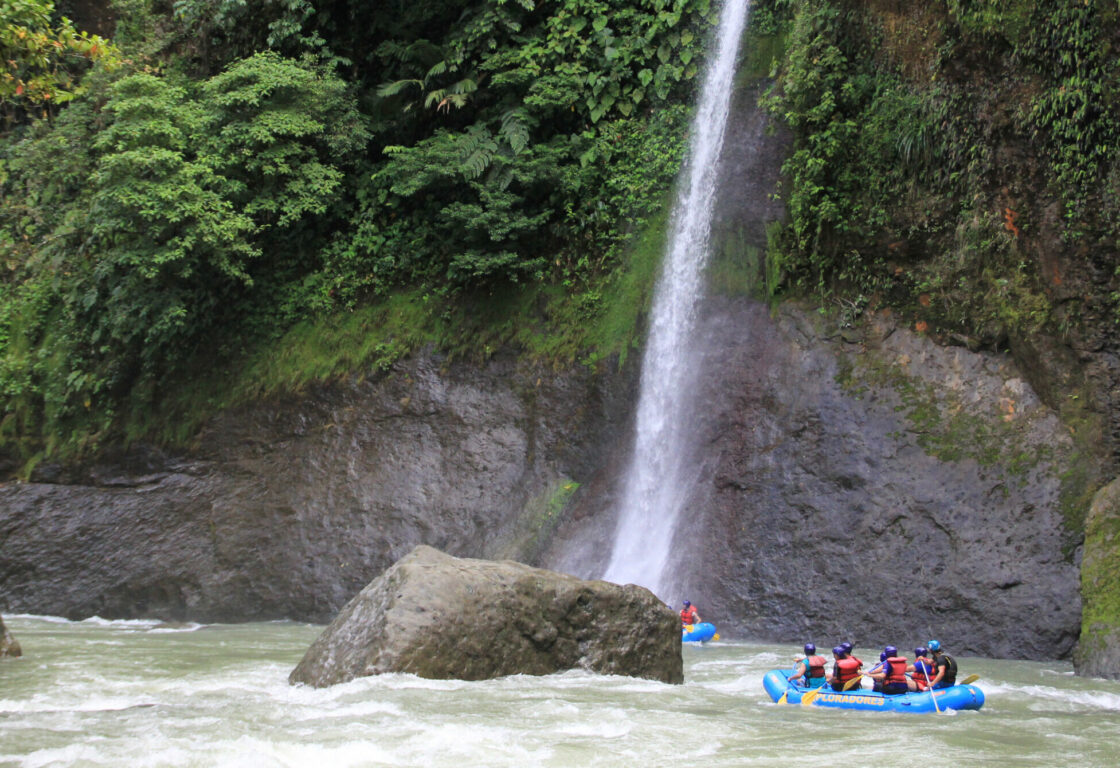 A group of tourists wearing lifejackets in a white water raft, in a rough looking river. Surrounding the river are boulders fringed with rainforest trees and a long waterfall pooling into the river.