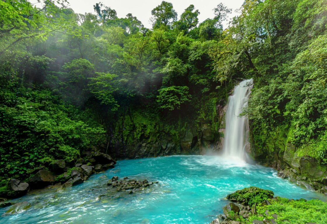 Tuquoise blue natural pond with waterfall flowing into it, Lush green forest surrounding.