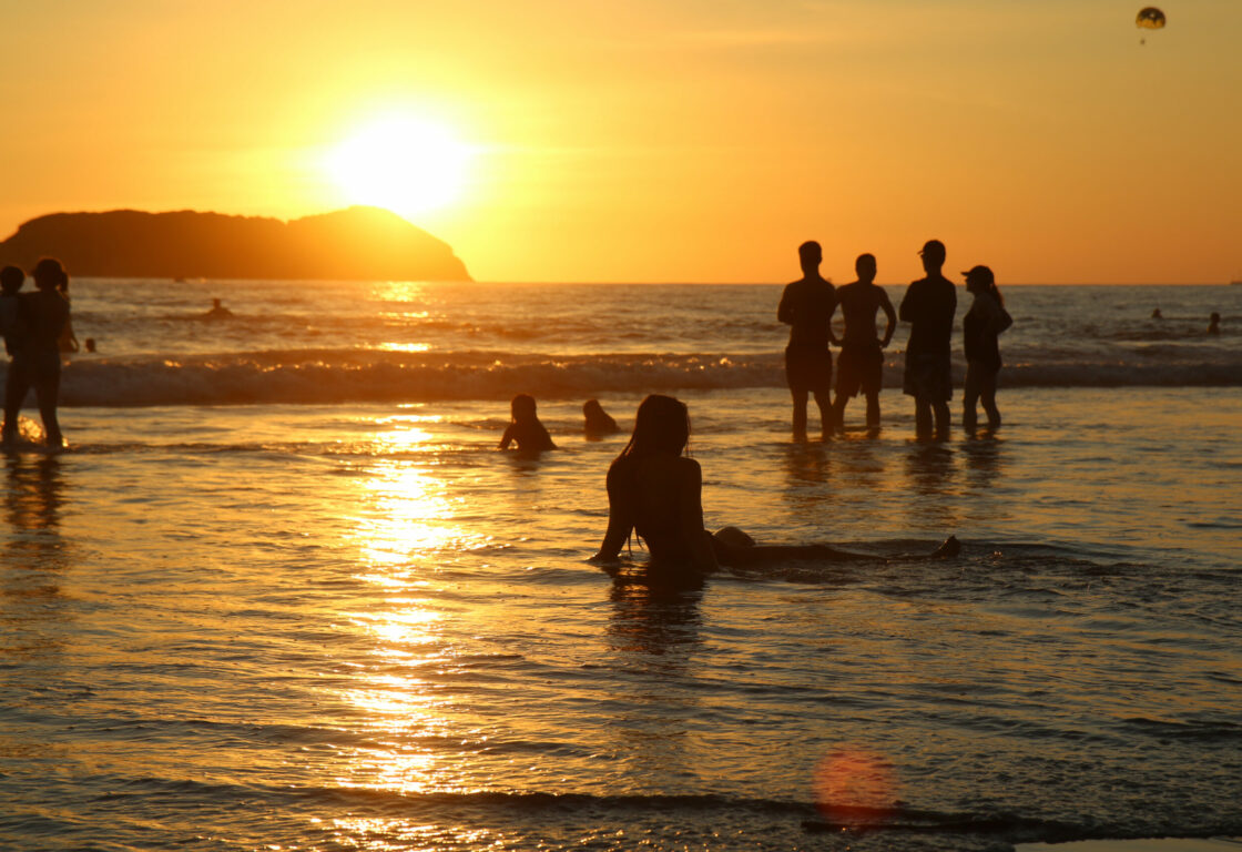 Groups of people stand and lie about in the sea, while the sun sets on the horizon