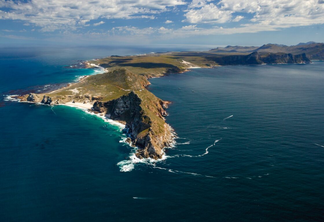 Aerial view of a rugged, green coastline extending into a vast, deep blue ocean under a partly cloudy sky, conveying a sense of natural beauty and remote tranquility.