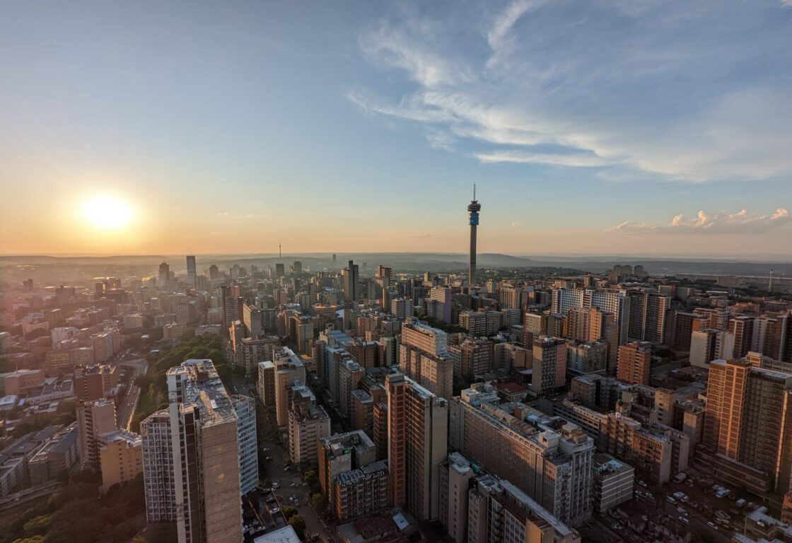 Aerial view of a city skyline at sunset, with tall buildings and a prominent tower under a partly cloudy sky, evoking a serene, golden ambiance.