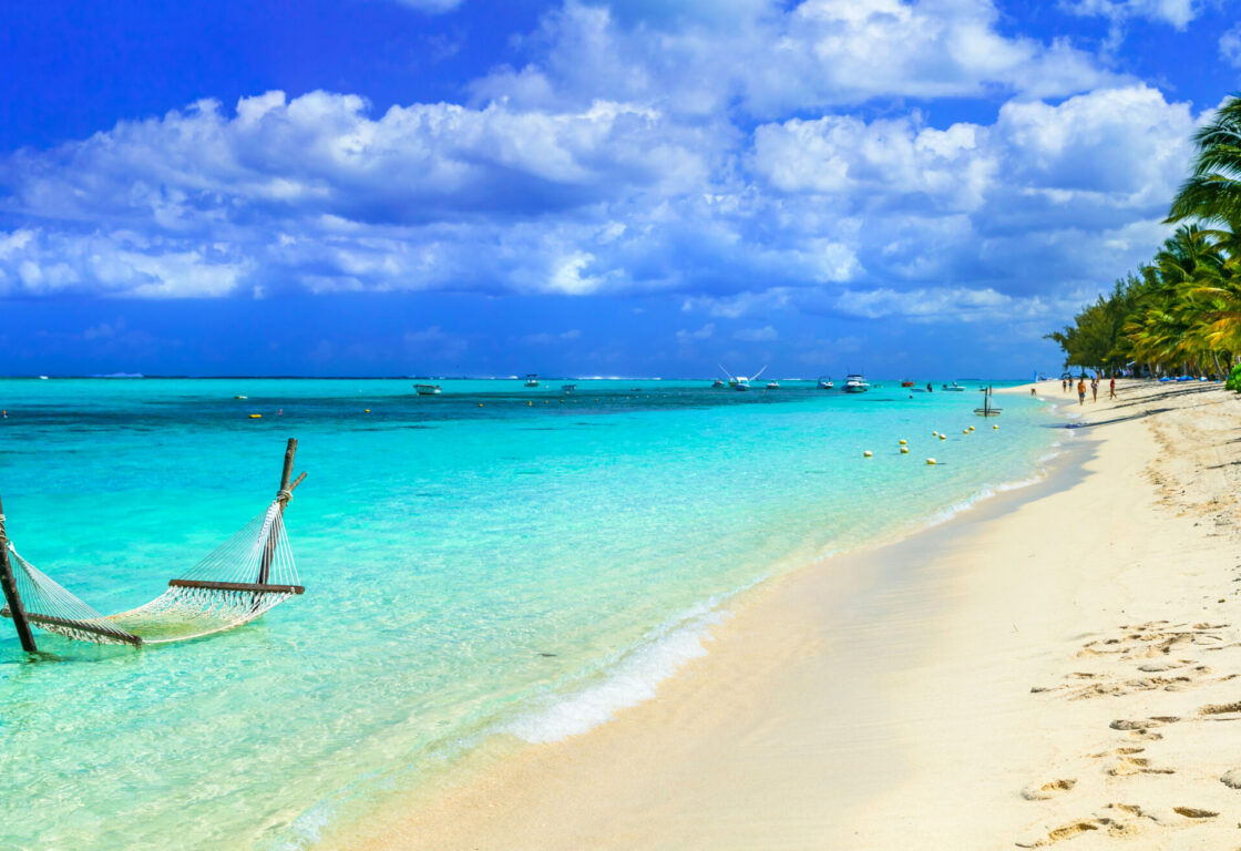 Tropical beach scene with a hammock in turquoise water, golden sand, and palm trees under a vibrant blue sky. Boats dot the distant horizon.
