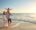 A family of four enjoys a sunny beach walk. The parents, wearing casual clothes, hold two young children. One child sits on a shoulder, the other held closely. Waves crash gently nearby.
