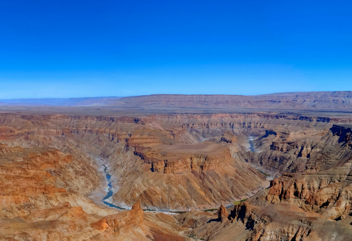 Panoramic view of Fish River Canyon under a clear blue sky. The rugged, red rock formations create a dramatic, expansive landscape.