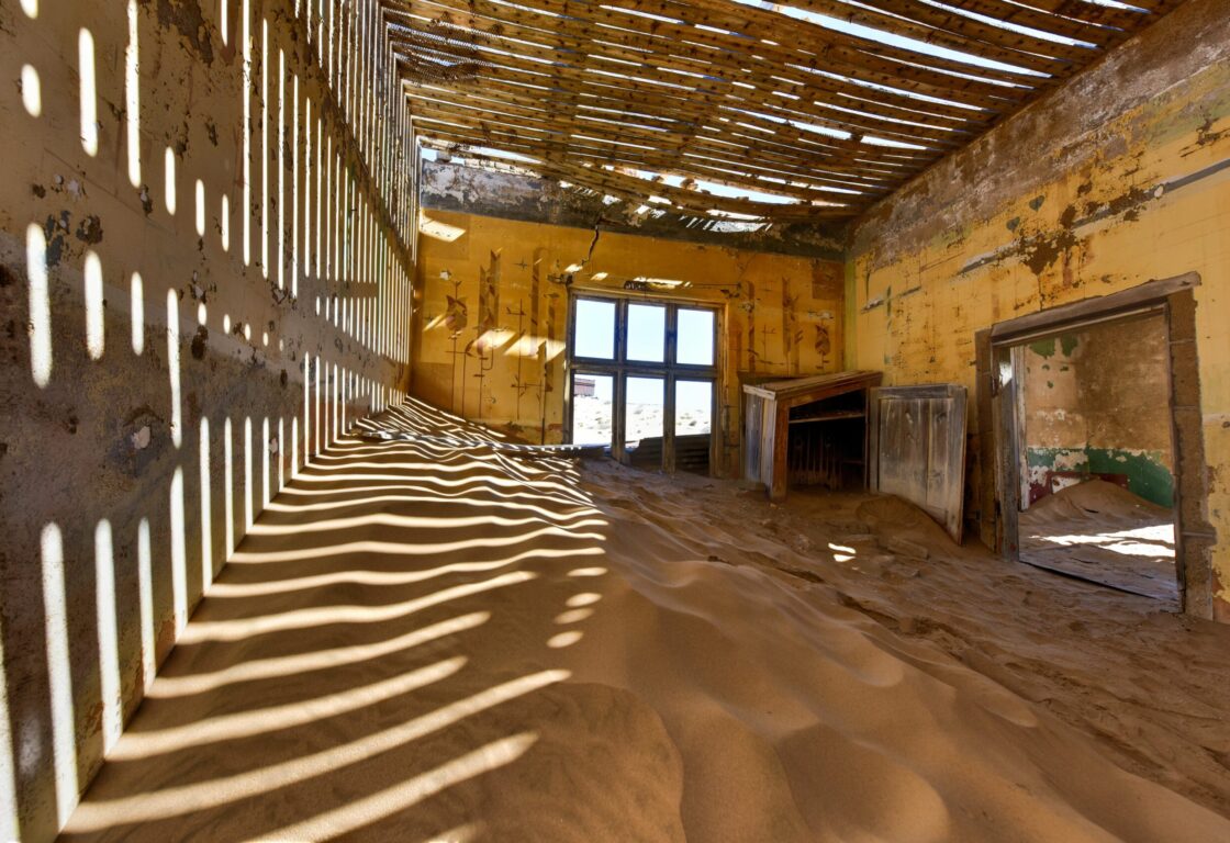Abandoned room filled with sand, sunlit through weathered wooden slats above. The walls are cracked and peeling, creating a haunting, deserted ambiance.