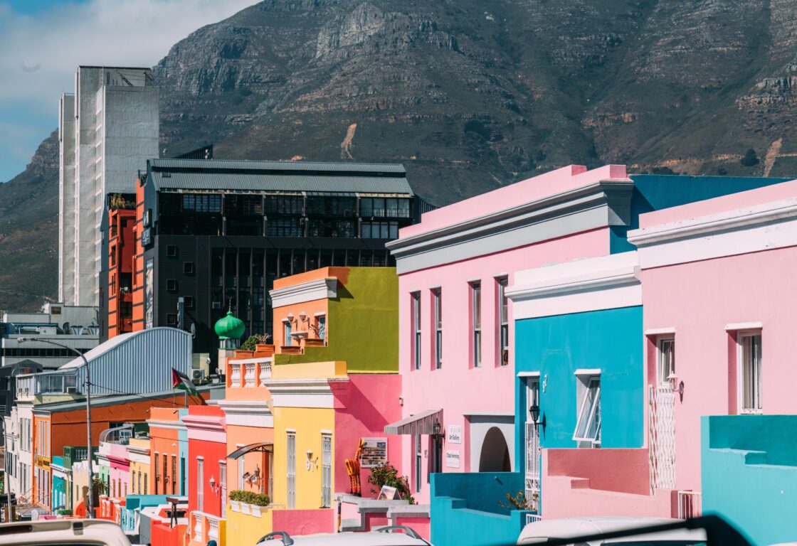 Colorful Bo-Kaap houses with pink, blue, yellow, and green facades line the street. Mountain backdrop and modern buildings contrast the vibrant scene.