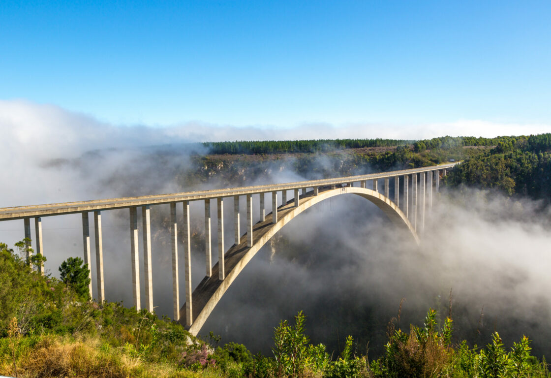 A long concrete arch bridge spans a misty valley, surrounded by lush green forest. The scene is serene, with the bridge emerging through low-hanging clouds.