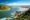 Aerial view of coastal lagoon on South Africa’s Garden Route with turquoise water, sandy estuary, green hills, rocky coastline, and distant mountains beneath a bright blue sky.