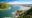Aerial view of coastal lagoon on South Africa’s Garden Route with turquoise water, sandy estuary, green hills, rocky coastline, and distant mountains beneath a bright blue sky.