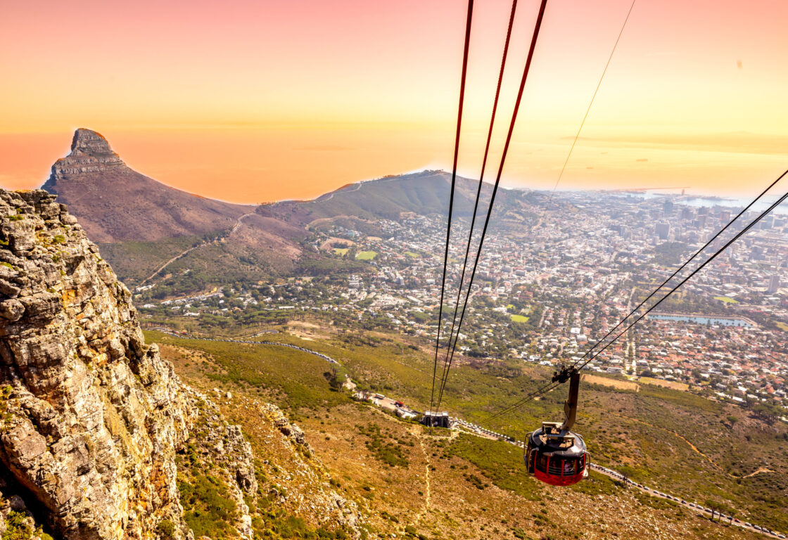 Cable car ascending Table Mountain with a panoramic view of Cape Town at sunset. Lion's Head peak on the left, cityscape sprawling below.