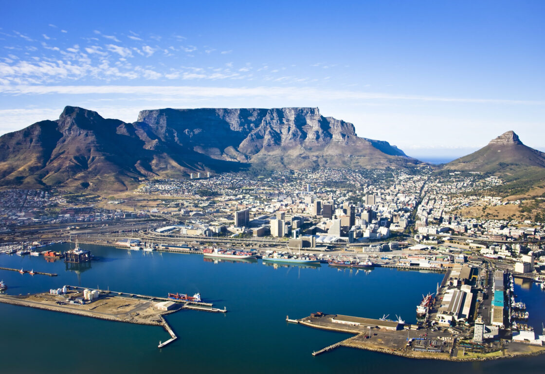 Aerial view of Cape Town with iconic Table Mountain in the background, blue sky above, cityscape and harbor in foreground, conveying a sense of vibrant beauty.