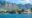 A vibrant coastal scene with two people kayaking on clear blue water, set against rocky shores and lush greenery. Majestic mountains rise in the background.
