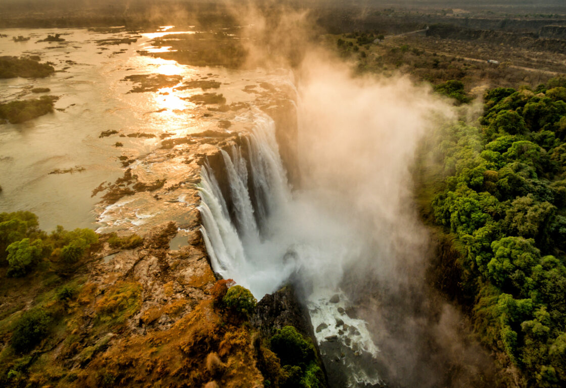 Aerial view of a majestic waterfall with a massive curtain of white frothy water cascading off a cliff. At the bottom, mist is rising, surrounded by lush green forest. The sun sets in the background, casting a golden glow.