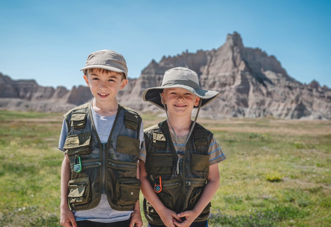 Two smiling boys in adventure vests and hats stand in a grassy field with rugged, sunlit mountains in the background under a clear blue sky.
