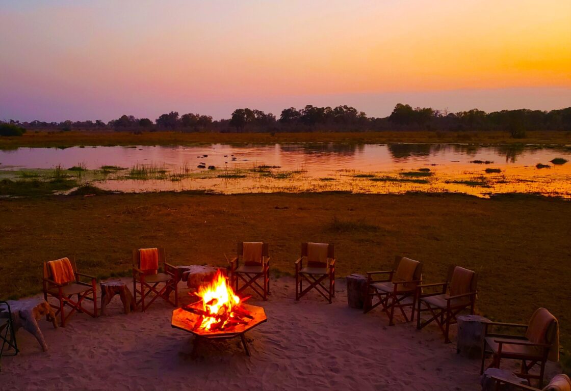 Campfire at sunset with around 10 canvas chairs placed around in a circle. In the background is a river, lit up orange from the reflection of the setting sun.
