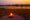 Campfire at sunset with around 10 canvas chairs placed around in a circle. In the background is a river, lit up orange from the reflection of the setting sun.