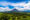 View of a conical volcano surrounded by lush tropical greenery under a blue and white cloudy sky