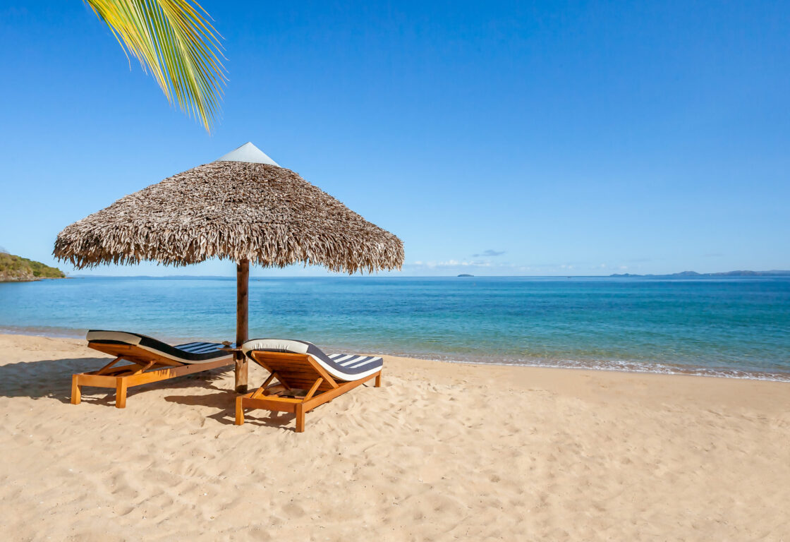 two wooden lounge chairs with blue and white stripy cushions on a Nosy Be beach with straw style umbrella