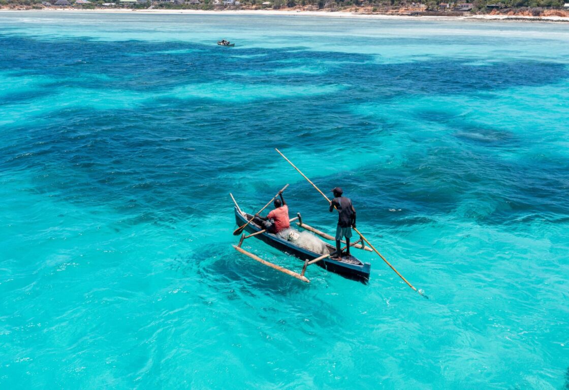 A small narrow row boat with two fishermen in it, floats on the completely still tropical Mozambique sea. The water is light turquoise and completely clear, revealing coral reefs below the water and small fish.