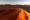 footprints on the peak of a sand dune, the sun rises, casting an orange glow on the sand.