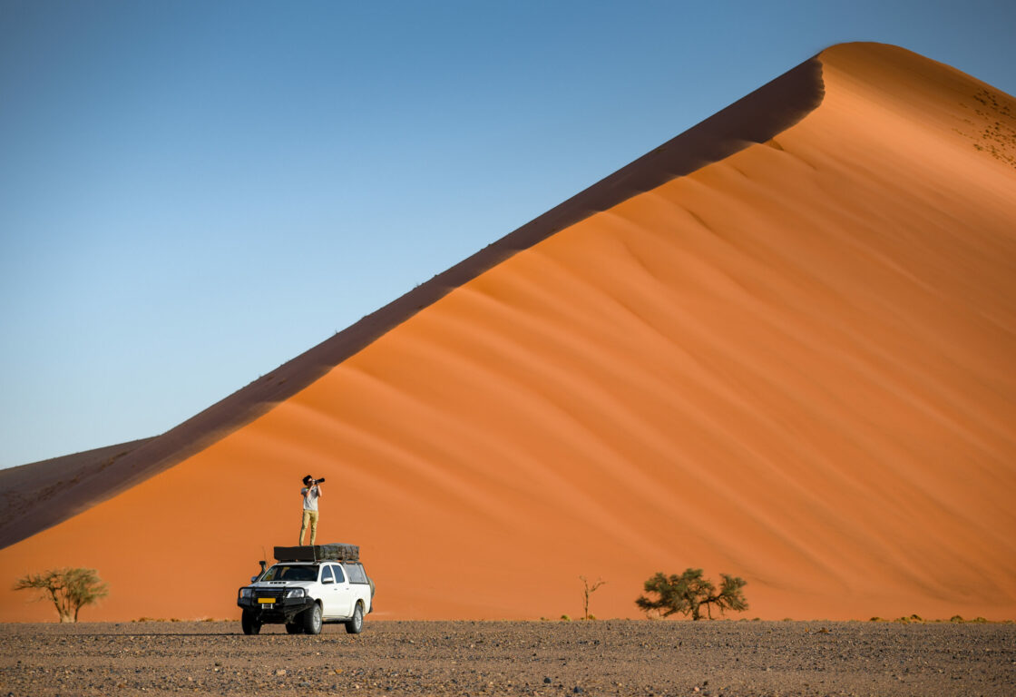 Young man standing on camper car near orange sand dune 45 taking photos.