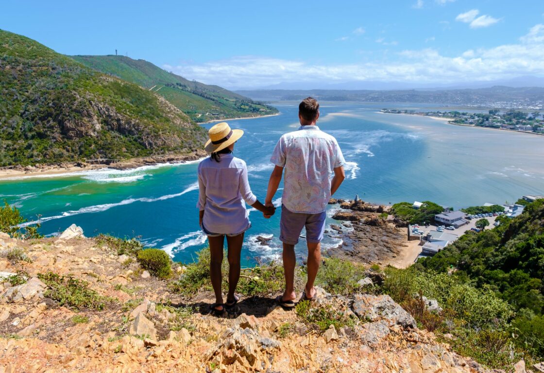 a honeymoon couple standing atop a hill looking down at stunning sea scenery with green hills in the background - Knysna, South Africa