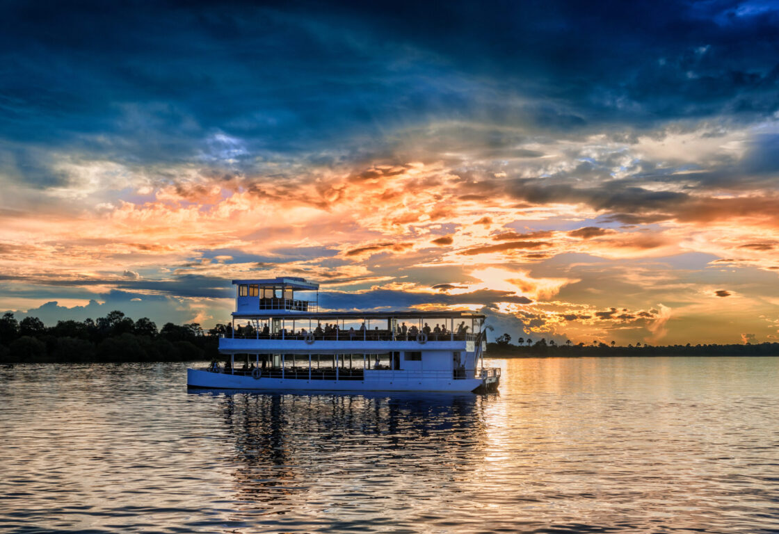 A white double deckered river boat crowded with people glides through the calm waters of a river whilst overhead the sun sets against a cloudy sky.