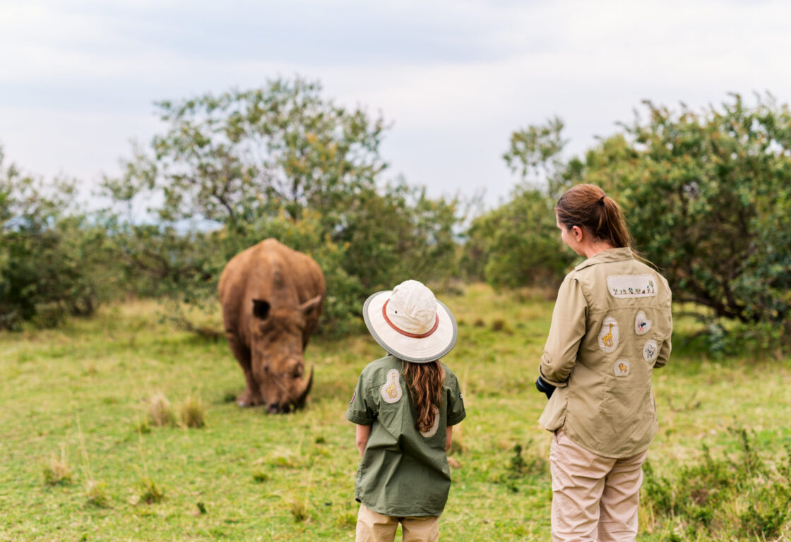 a small girl and a safari guide watch a rhino grazing on grass during a safari holiday