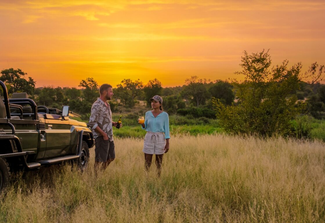 a couple having sundowner drinks by a safari jeep in the savanah at sunset