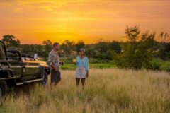 a couple having sundowner drinks by a safari jeep in the savanah at sunset