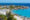 A scenic beach in Cape Town with white sand, turquoise-blue water, and large granite boulders. People relax under blue umbrellas, swim in the calm sea, and paddleboard near the shore. Palm trees and coastal houses line the backdrop under a clear blue sky.