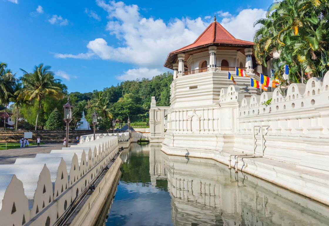 Exterior view of the Temple of the Tooth in Kandy, Sri Lanka, with its white walls, red roof, and reflecting moat under a blue sky.