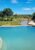 A serene infinity pool overlooks a dry riverbed and lush green bushveld under a clear blue sky. A wooden deck with lounge chairs sits beside the pool, offering an elevated view across the tranquil, tree-dotted landscape.