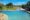 A serene infinity pool overlooks a dry riverbed and lush green bushveld under a clear blue sky. A wooden deck with lounge chairs sits beside the pool, offering an elevated view across the tranquil, tree-dotted landscape.