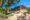 The outdoor common area of a safari lodge featuring a large circular thatched-roof building with dry-stone walls, stone steps leading up to an open veranda, flagstone terracing with metal bistro tables and chairs, and mature trees providing dappled shade under a vivid blue sky