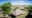 An elevated view of two thatched-roof safari tent structures on raised platforms beside a large acacia tree, set within open grassland on the edge of the vast Serengeti plains under a bright blue sky.