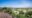 A view from atop a granite kopje looking down over two thatched safari camp structures nestled among lush green acacia trees, with the vast open Serengeti plains stretching to the horizon under a bright blue sky.