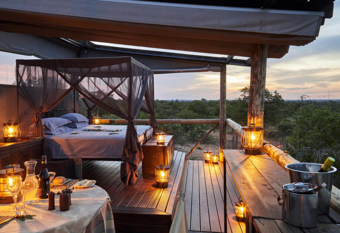 Photo is shot at dusk with beautiful light and candled lanterns scattered around a covered wooden sleepout deck with green scrubland running away into the distance as the sun sets in a cloudy sky. A linen covered table laid for dinner and nice bucket with wine sit in the foreground and four poster bed with mosquito nets draped around it in the background complete the romantic scene.