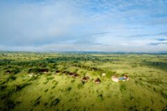 A high aerial view of a safari camp with a string of thatched circular structures and a turquoise swimming pool, perched along a lush green ridge overlooking the vast rolling Serengeti plains, with the winding Mara River visible in the far distance
