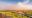 A lone deck chair and small side table on a weathered hardwood viewing deck with a rustic pole railing, overlooking a breathtaking panorama of the Mara River winding through vivid green plains, under a vast dramatic sky of rose-hued dust haze and blue