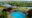 An aerial view of a circular swimming pool on a curved hardwood deck with sun loungers, a thatched pool bar pavilion to one side, positioned dramatically on a clifftop directly above the wide, winding Mara River with lush green plains beyond