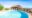 A large curved swimming pool on a stone terrace with a thatched open-sided pool bar and guests relaxing on wooden sun loungers, set against a sweeping panorama of the Serengeti plains and distant escarpment under a deep blue sky