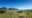 A row of five thatched safari lodge structures with raised wooden decks, positioned along a lush green hillside crest overlooking the vast open Serengeti plains and a distant ridge, under a deep cloudless blue sky