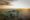 An aerial drone view of a small thatched-roof safari camp nestled among acacia trees and a large granite boulder on the open Serengeti plains at golden hour, with sunlight streaming across the vast landscape to the horizon.