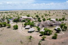 An aerial view of a small luxury safari camp with thatched accommodation units and a turquoise swimming pool, built around large granite kopje boulders and connected by stone-edged pathways, set within the boundless expanse of the Serengeti plains.