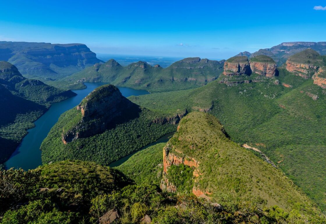 An aerial view of three massive round mountain peaks rising above a deep green canyon, surrounded by winding rivers, steep cliffs, and endless stretches of rugged natural landscape.
