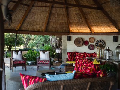 Chundukwa River Lodge an open space with bamboo roof and wicket furniture scattered with cushions and on one white wall there are plates decorated with different art and the other side opens onto green trees and grass area
